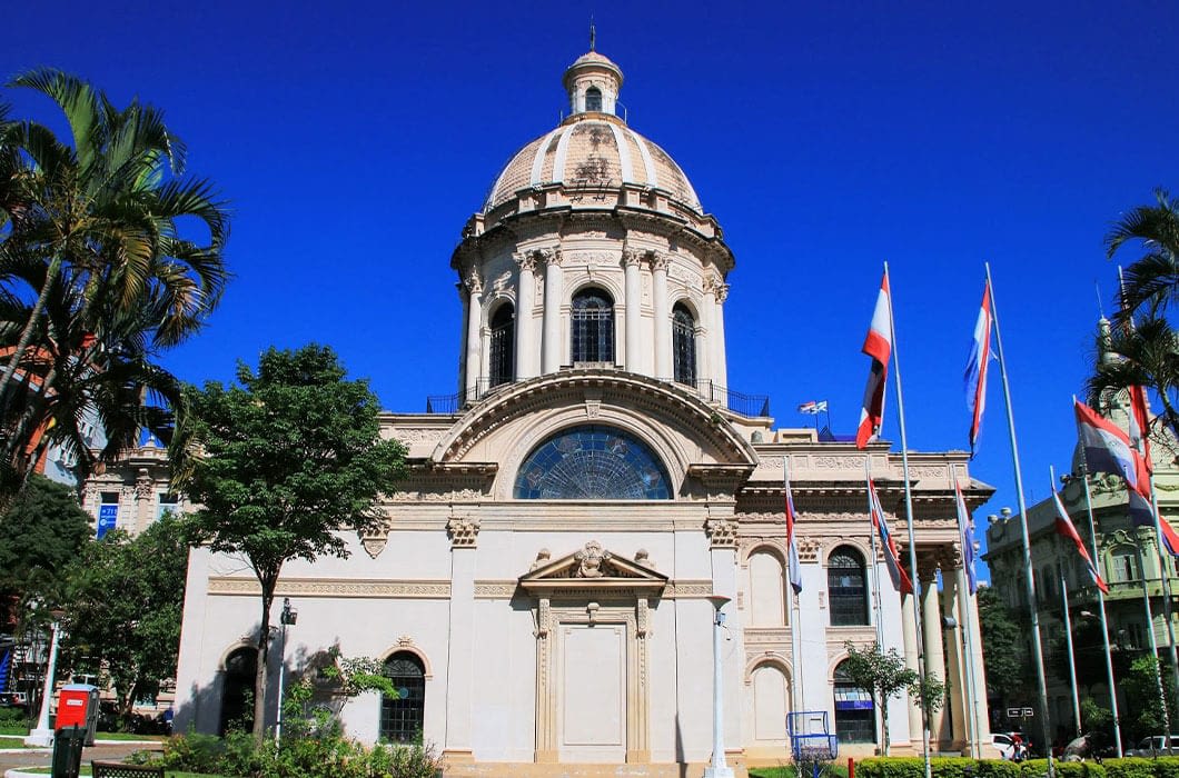 A grand domed building with classical architecture, tall arched windows, palm trees, and several national flags in front under a clear blue sky—one of the top Paraguay attractions for those planning Paraguay travel.