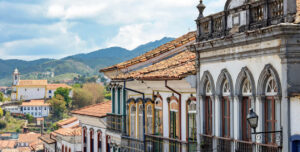 Colorful colonial buildings with arched windows and balconies line a street—a scene that showcases some of the amazing places in Brazil, with a church and green hills visible in the background under a partly cloudy sky.