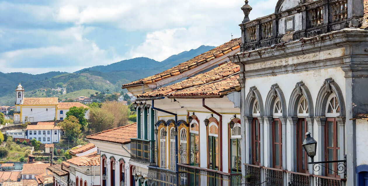 Colorful colonial buildings with arched windows and balconies line a street—a scene that showcases some of the amazing places in Brazil, with a church and green hills visible in the background under a partly cloudy sky.