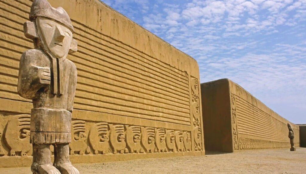 Large ancient adobe wall with intricate carvings and two tall stone statues in front, under a partly cloudy sky—an archaeological site possibly linked to Pachacuti, the legendary founder of the Incan Empire.