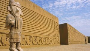 Large ancient adobe wall with intricate carvings and two tall stone statues in front, under a partly cloudy sky—an archaeological site possibly linked to Pachacuti, the legendary founder of the Incan Empire.