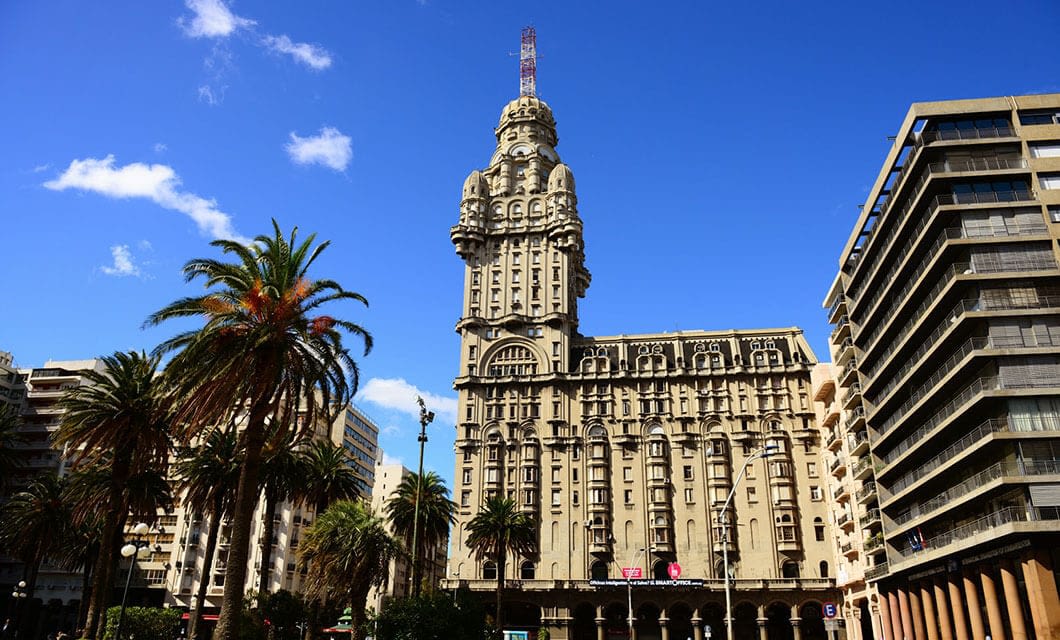 A historic, ornate building with a tall central tower rises amid palm trees and modern buildings under a bright blue sky—one of the must-see Uruguay attractions for those planning Uruguay travel.