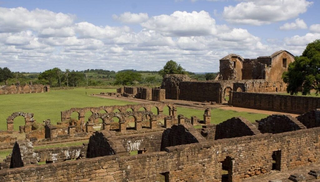 Ancient stone ruins of a large building complex stand on grassy land under a partly cloudy sky, with arches and walls visible—an evocative scene often explored on South America and Bolivia tours, surrounded by green trees and open fields.