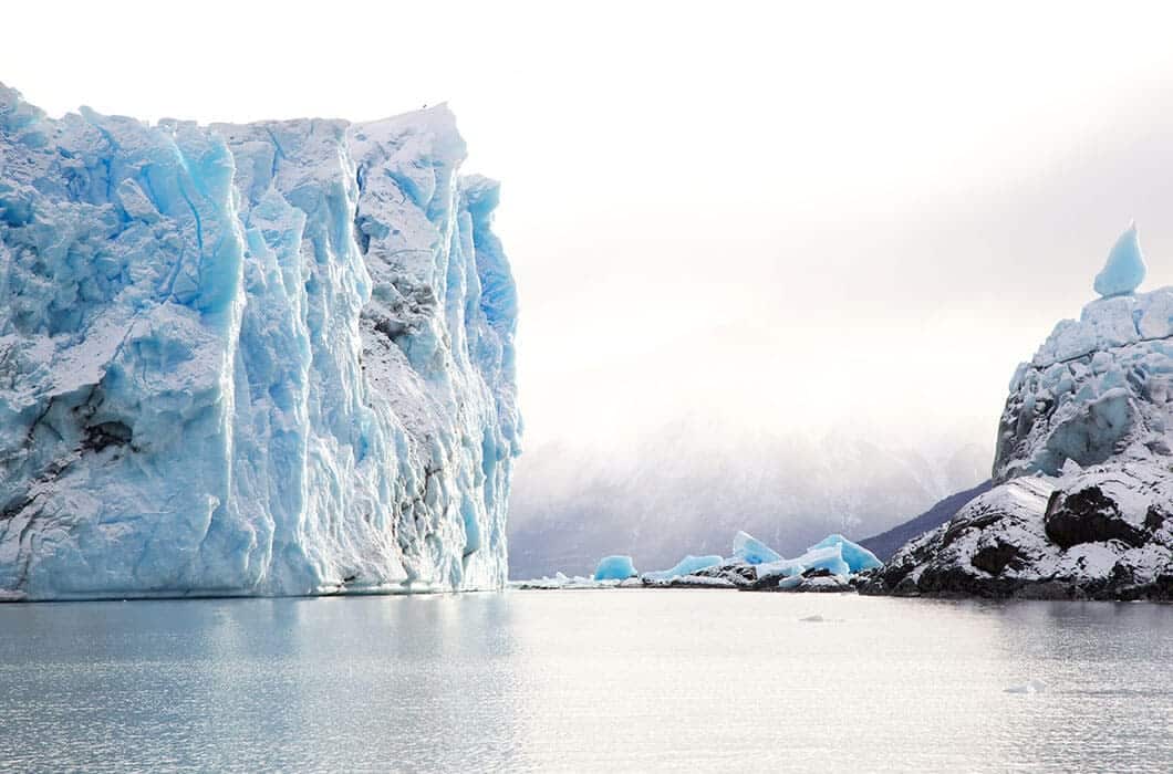 A large blue glacier with sharp, jagged edges rises from calm, icy water—one of the unforgettable things to do in Patagonia. Snow-covered mountains stand in the background beneath a cloudy, overcast sky.