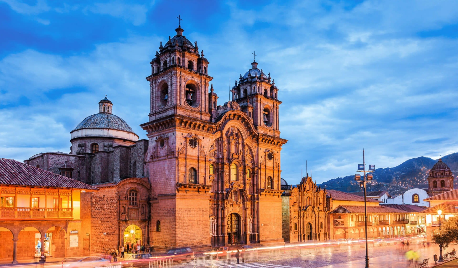 Historic stone cathedral with twin bell towers in Cusco, Peru, illuminated at dusk—a must-see among Peru attractions. The blue sky and blurred lights of passing cars create a magical scene, with mountains framing this top spot for Peru travel.