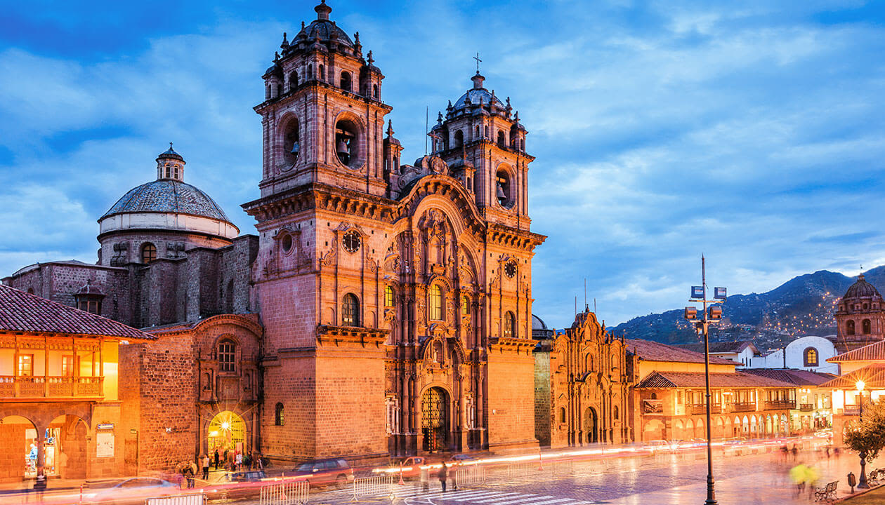 A historic stone cathedral with two bell towers stands illuminated at dusk in a lively city square in Peru, with blurred lights from passing vehicles and mountains visible in the background—a must-see for travel enthusiasts seeking unique places to visit.