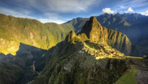 Ancient stone ruins of Machu Picchu sit atop a lush green mountain ridge in Peru, surrounded by steep forested peaks under a bright blue sky. Sunlight casts dramatic shadows across the breathtaking landscape.
