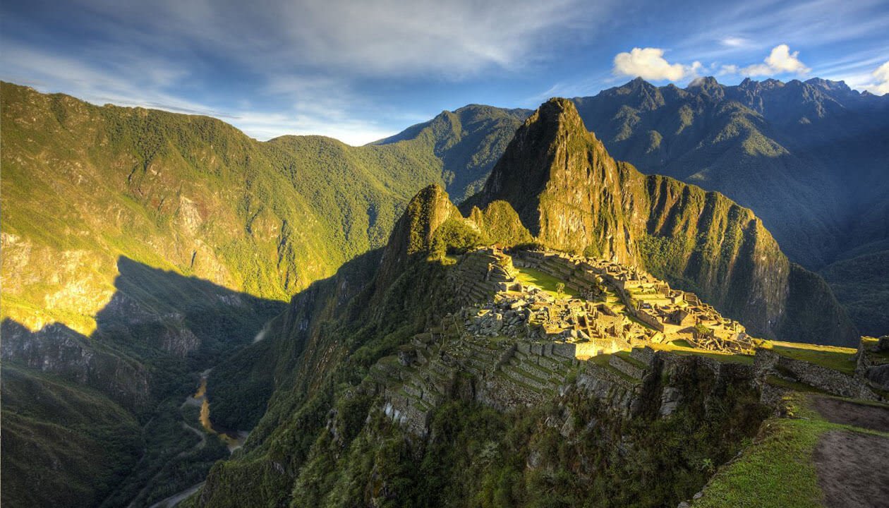 Ancient stone ruins of Machu Picchu sit atop a lush green mountain ridge in Peru, surrounded by steep forested peaks under a bright blue sky. Sunlight casts dramatic shadows across the breathtaking landscape.