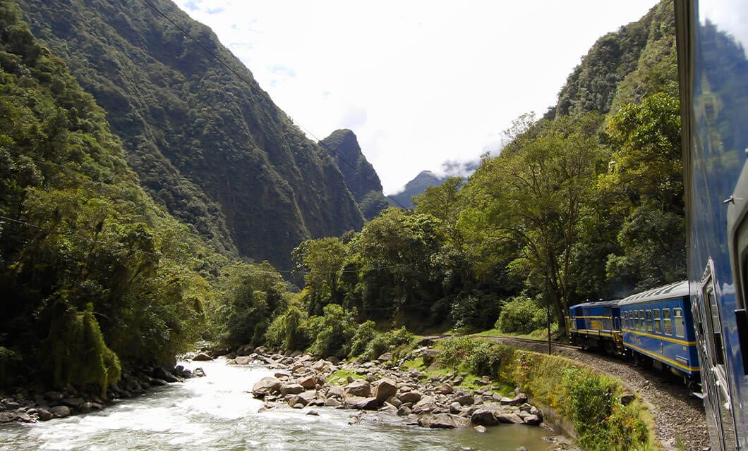 A blue and yellow train travels alongside a river, winding through lush green mountains under a partly cloudy sky—a breathtaking view among top Peru attractions for unforgettable Peru travel. Dense trees and steep cliffs surround the scene.