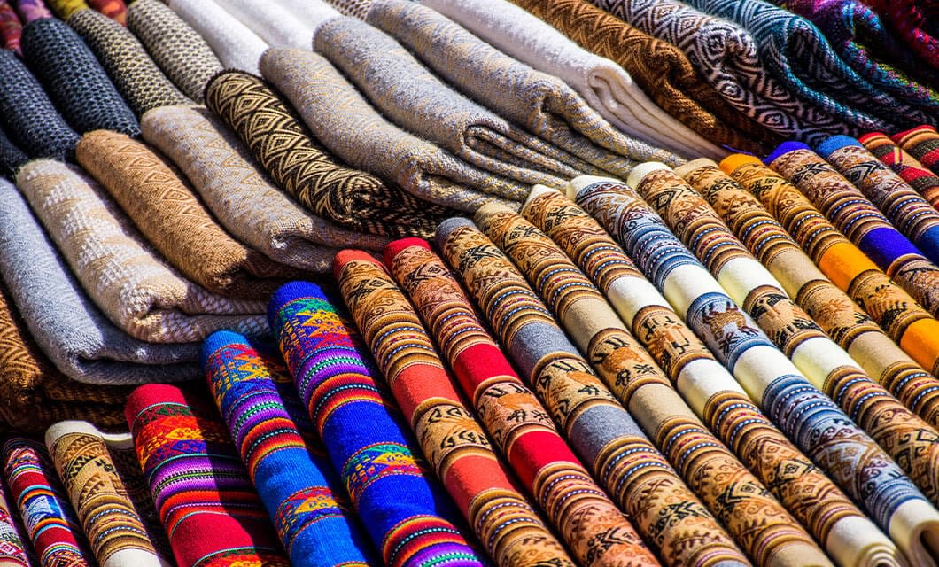 Stacks of colorful, patterned woven fabrics and blankets are neatly arranged, showcasing intricate designs and a variety of bright and earthy tones—a true highlight among Peru attractions for those interested in traditional craftsmanship.
