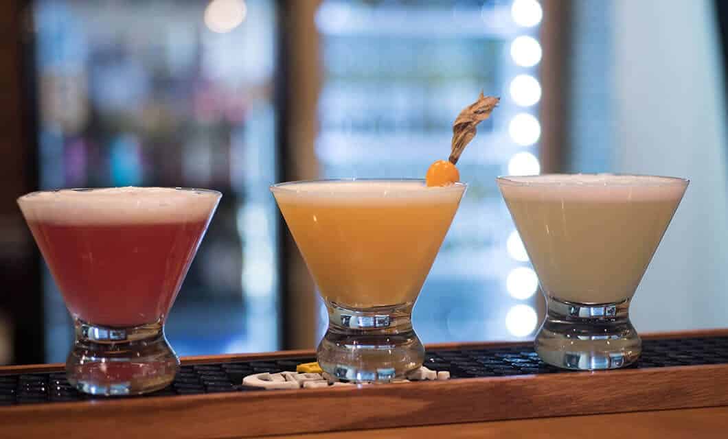 Three colorful cocktails in stemmed glasses sit on a bar; the drinks—red, yellow with a garnish, and pale green—all topped with foam, offer a taste of Peru. A softly blurred background showcases lights and shelves—a must-see for travel enthusiasts.