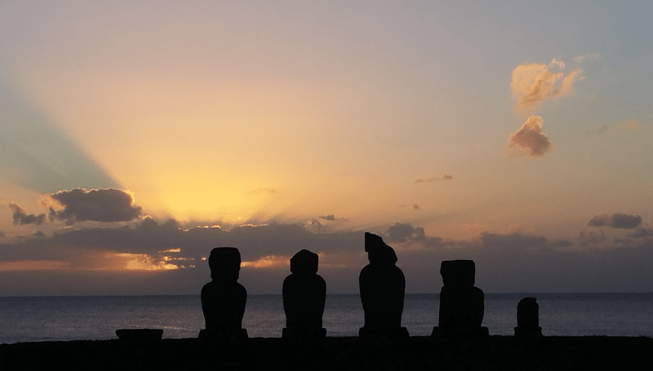 Four moai statues are silhouetted against a colorful sunset sky on Easter Island, with rays of sunlight streaming through clouds over the ocean in the background.