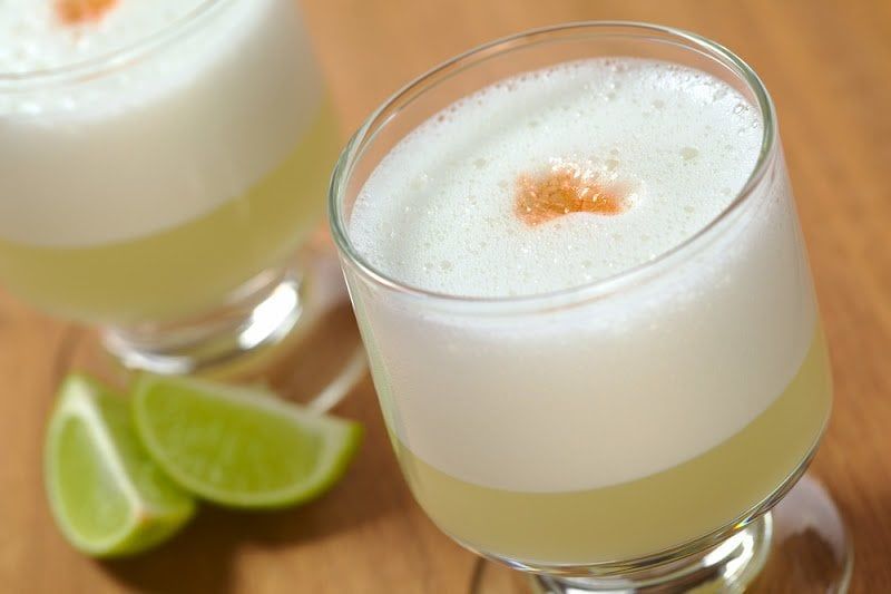 Two glasses of frothy pisco sour cocktail, a classic among South American drinks, each topped with a dash of bitters, sit on a wooden surface next to two lime wedges.