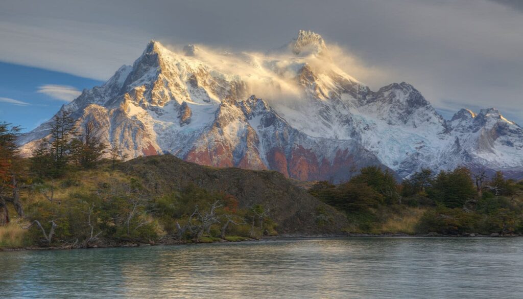 Snow-capped mountains rise above a calm lake near Puerto Natales, with sunlight illuminating the peaks. Rocky slopes and patches of autumn trees frame the foreground—perfect for travel photography and discovering new things to do.