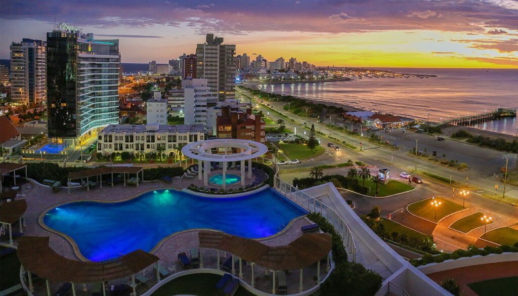 Aerial view of a city at sunset with a large illuminated pool in the foreground, modern high-rise buildings, coastal road, and the shoreline with a marina—an iconic scene for Uruguay travel and essential for any list of places to visit in Uruguay.