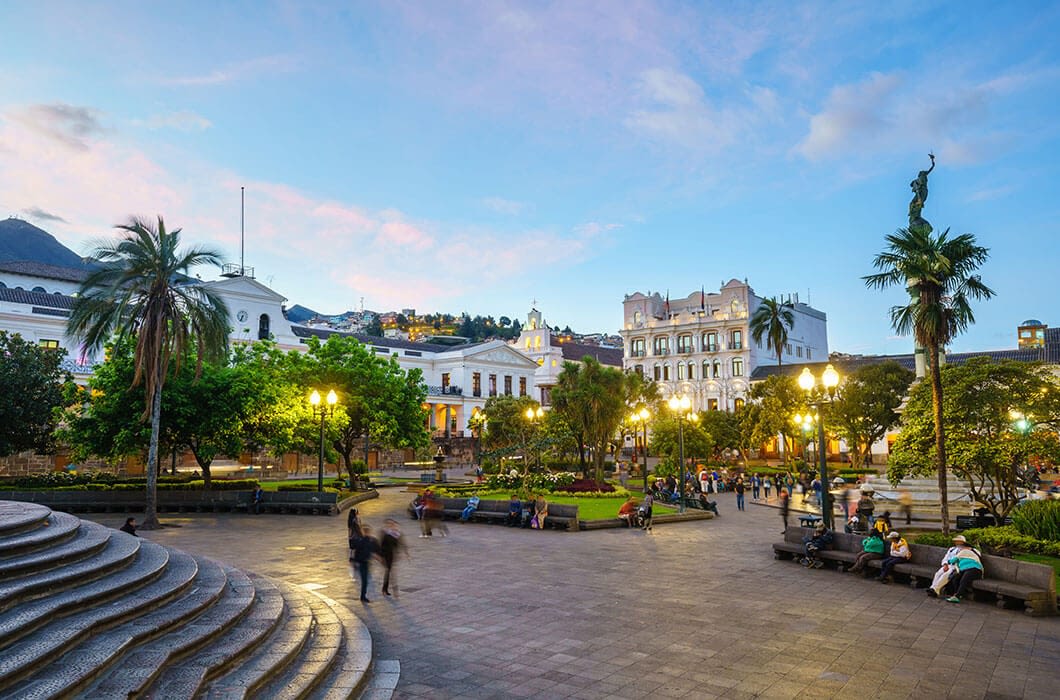 A lively public square with trees, benches, and people relaxing—one of the top Ecuador attractions. Surrounded by historic buildings under a blue sunset sky, lamp posts light the scene, with a statue and distant hills in the background.