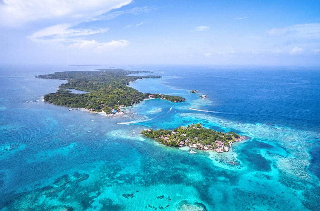 Aerial view of lush, green islands surrounded by vibrant turquoise and deep blue ocean water under a partly cloudy sky. Waves and reefs are visible, highlighting the tropical scenery and unforgettable Colombia travel experiences.