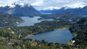 A scenic landscape from the Seven Lakes region in Patagonia, featuring a large lake surrounded by lush green forests, small islands, and snow-capped mountains in the background under a partly cloudy sky along the Grand Circuit.