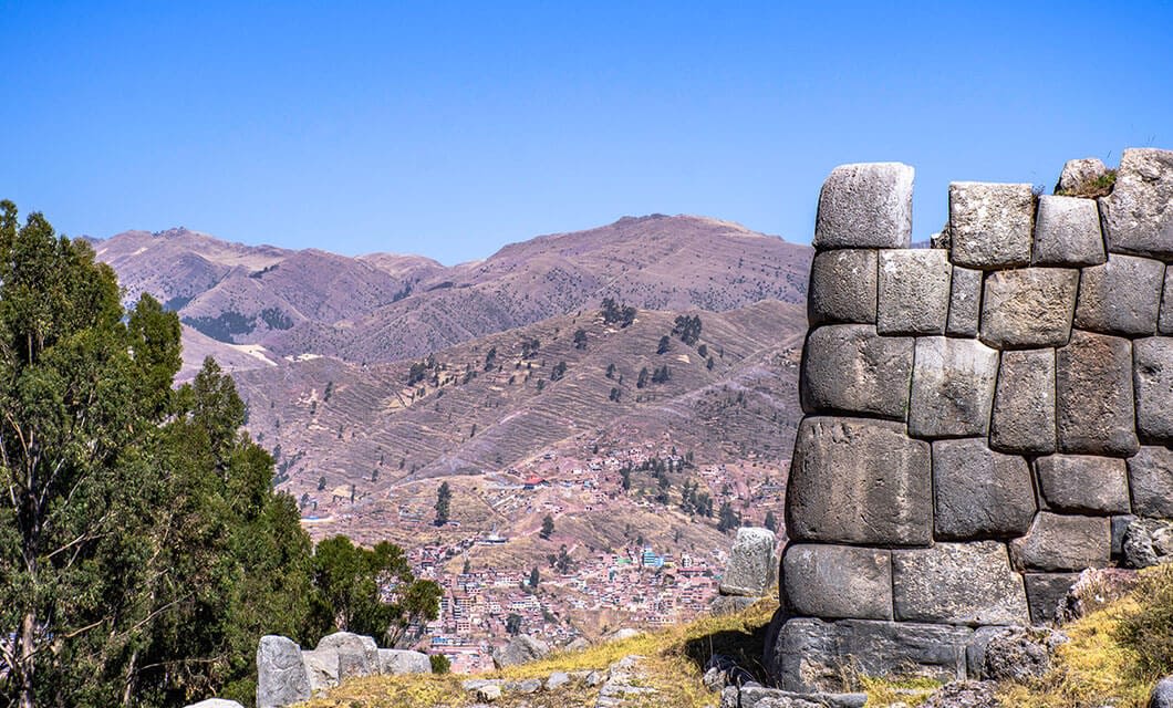 Ancient stone walls of Sacsayhuamán overlook Cusco and the mountains under a clear blue sky, with trees and terraced hills beyond—one of the must-see Peru attractions for anyone planning their Peru travel adventure.