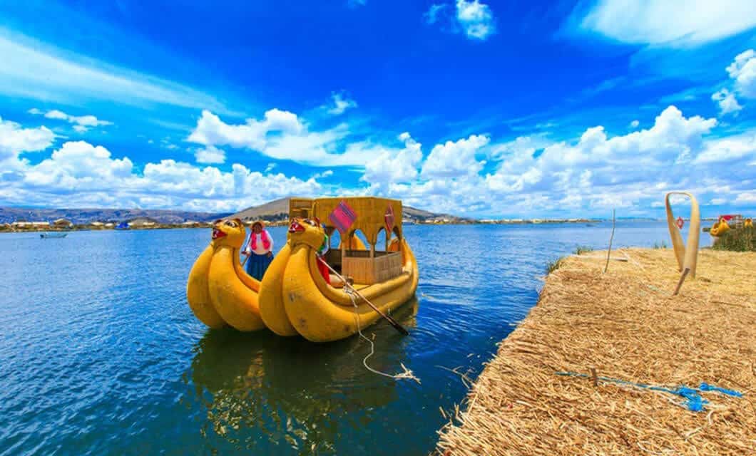 A traditional reed boat shaped like two yellow birds floats on a calm blue lake near a shore made of reeds, under a bright sky with scattered clouds—one of the unique Peru attractions for anyone seeking memorable Peru travel experiences.