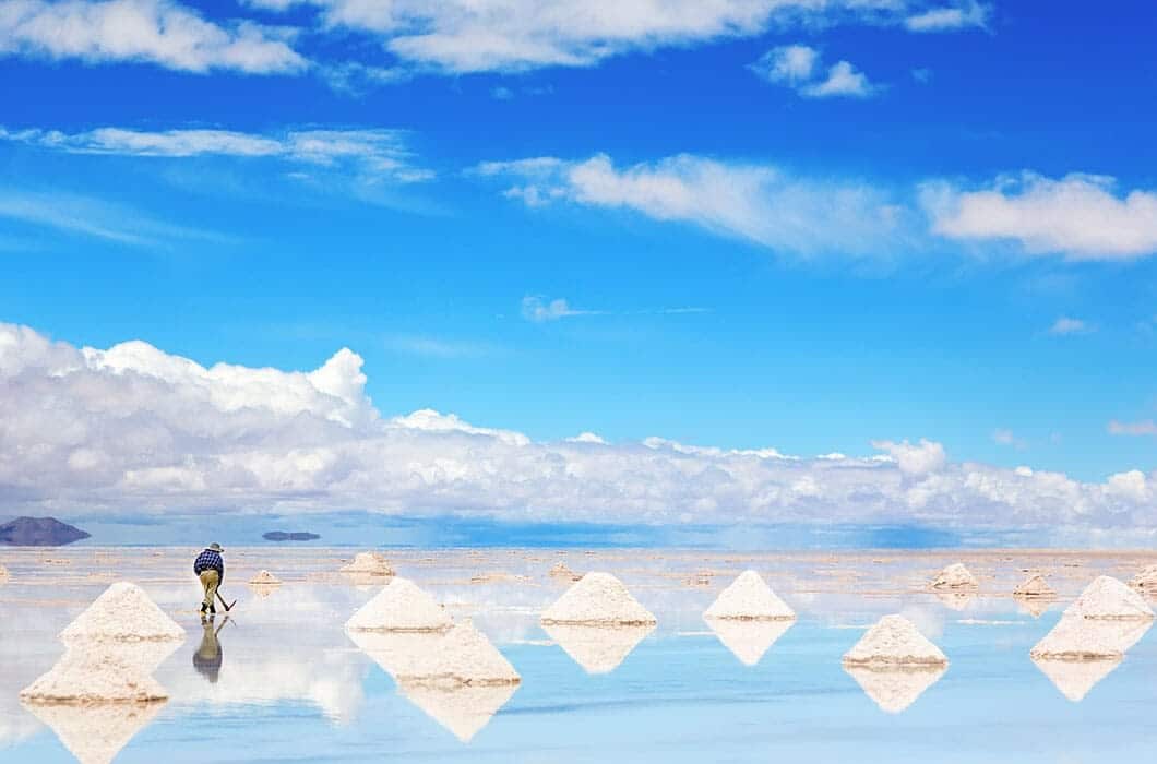 A person gathers salt among pyramid-shaped salt piles mirrored on a shallow surface beneath a bright blue sky. This serene, vast landscape is one of the top Bolivia attractions for those seeking unforgettable Bolivia travel experiences.