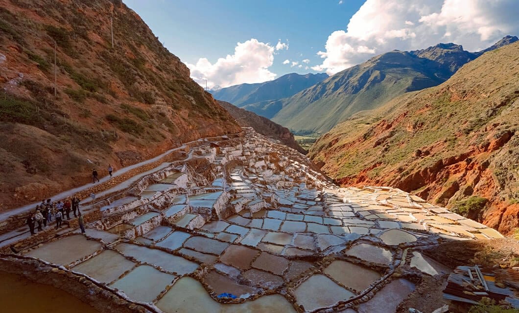 Terraced salt ponds nestle between red-brown hillsides, with mountains and clouds in the background. People walk near the lower left, showcasing one of the unique Peru attractions in this scenic valley landscape—a highlight for Peru travel.