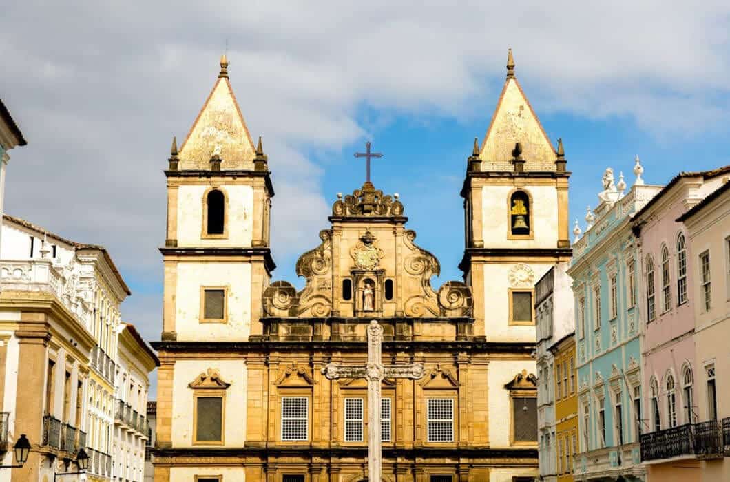 Historic baroque church with two bell towers and a cross on top, flanked by pastel-colored colonial buildings under a partly cloudy sky—a must-see among Brazil attractions for anyone exploring Brazil travel.