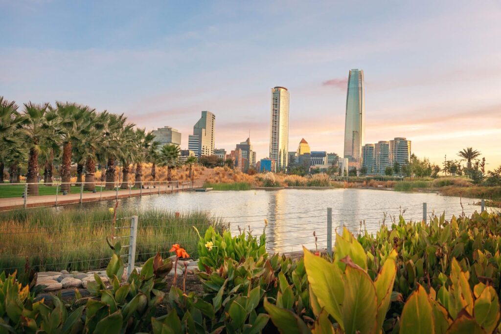 A city skyline with modern tall buildings behind a park of palm trees, lush green plants, and a large pond under a colorful sunset sky—a perfect scene for South America travel and discovering the best winter destinations.