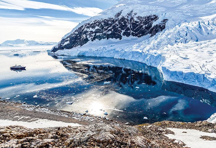 A glacier meets the calm, reflective water of the ocean in a polar landscape. Snow-covered mountains rise in the background, and a small boat floats near the ice edge—perfect for experiencing unique Antarctica activities under a partly cloudy sky.