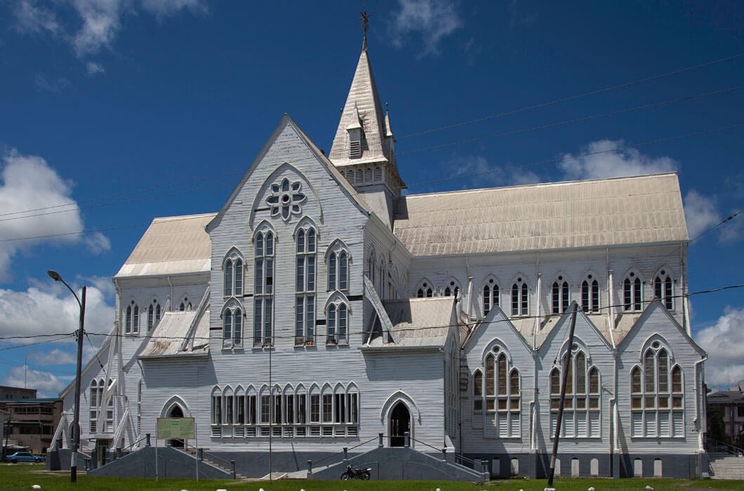 A large white wooden church with pointed arches and a tall steeple stands under a bright blue sky with scattered clouds—a must-see for those exploring the unique architecture among things to do in Guyana.