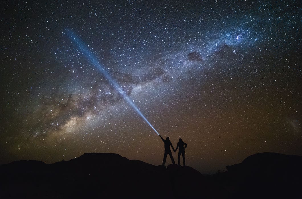 Two people stand on a hill under a starry night sky with the Milky Way visible; one shines a flashlight upward, creating a bright beam—an unforgettable moment for those exploring Things to do in Guyana or nearby French Guiana.