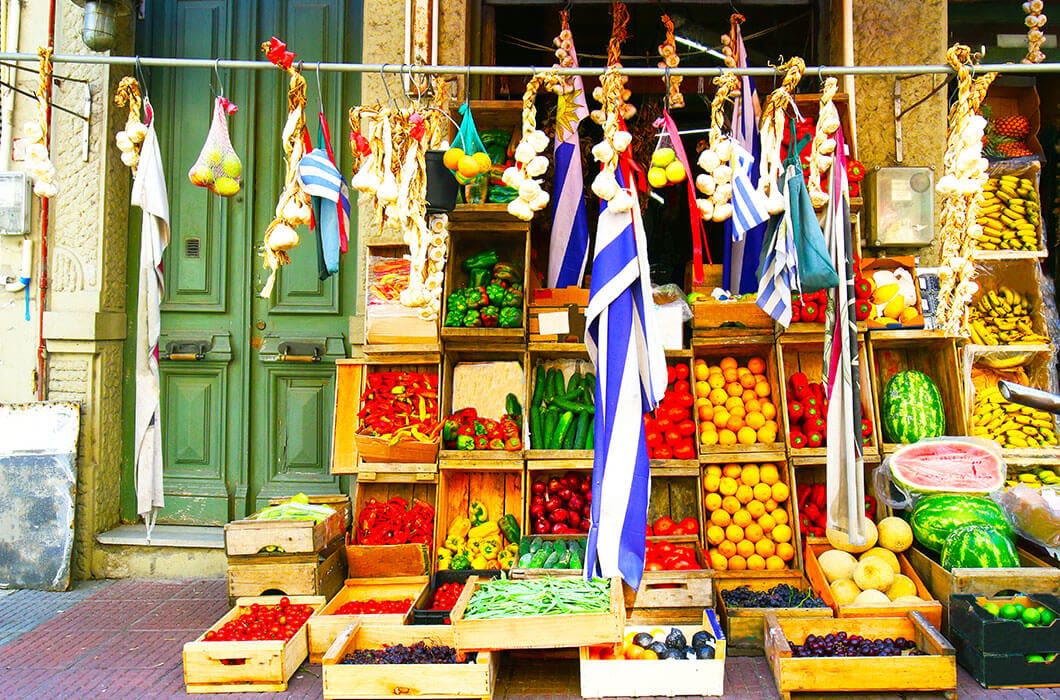 Colorful fruit and vegetable stand with wooden crates stacked full of produce, such as tomatoes, lemons, watermelons, bananas, and peppers—a lively scene reminiscent of Brazil attractions and popular things to do in Brazil. Two Greek flags are visible above.