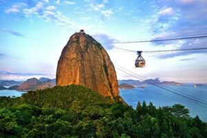 A cable car ascends to the top of Sugarloaf Mountain in Rio de Janeiro, Brazil, surrounded by lush greenery, blue ocean, and distant mountains under a partly cloudy sky.