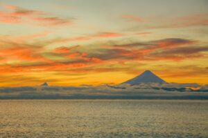 A serene sunset over calm water with two amazing volcanoes on the horizon, partially covered by clouds, the sky painted in warm orange, yellow, and pink hues—capturing the breathtaking beauty of South America.