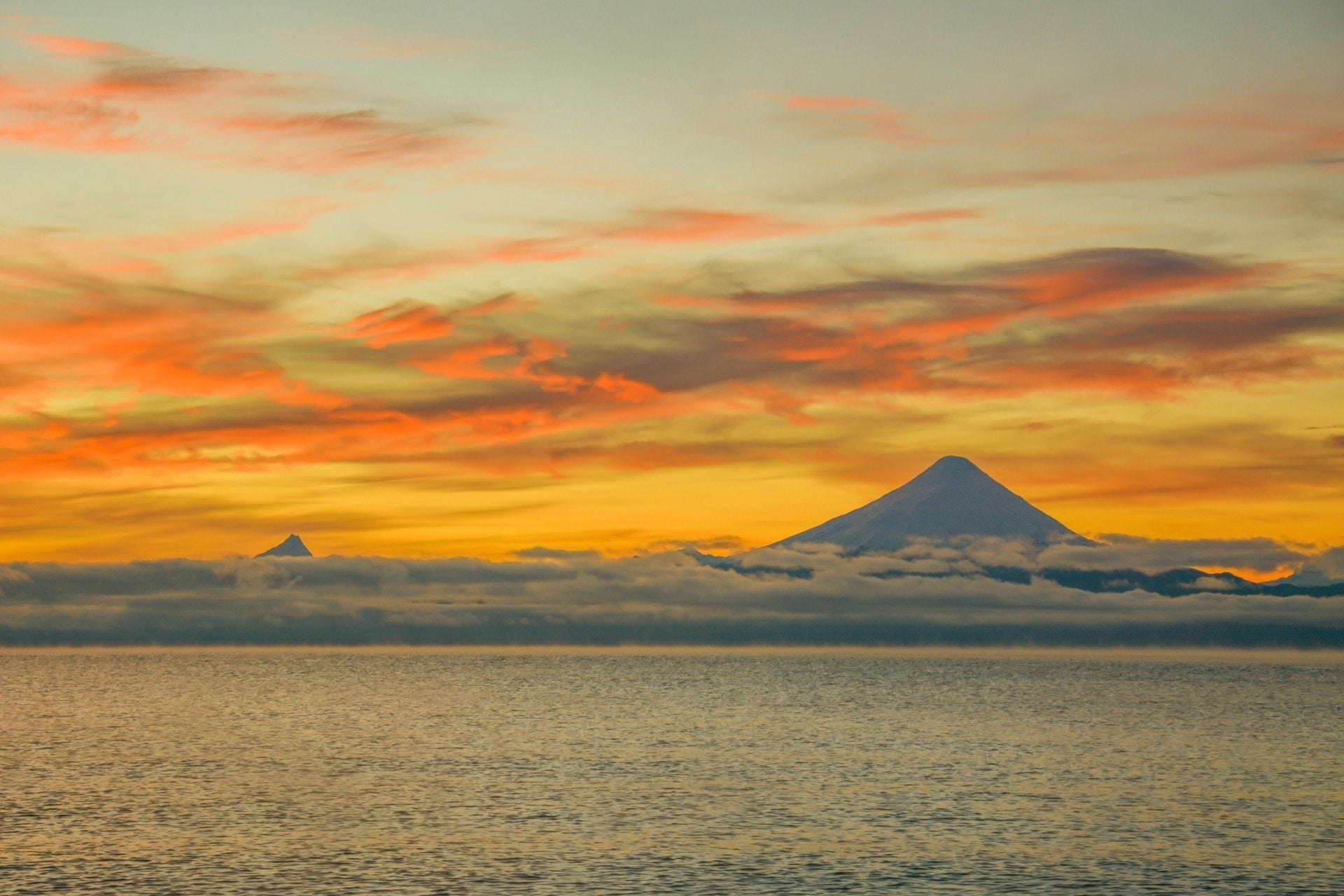 A serene sunset over calm water with two amazing volcanoes on the horizon, partially covered by clouds, the sky painted in warm orange, yellow, and pink hues—capturing the breathtaking beauty of South America.