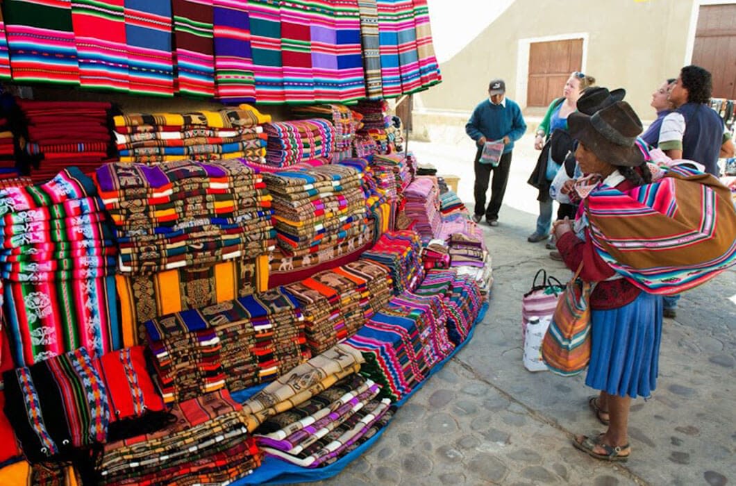 A woman in traditional clothing stands by a market stall displaying colorful woven textiles, while people browse on a cobblestone street—a vibrant scene among the many Bolivia attractions for those seeking authentic Bolivia travel experiences.