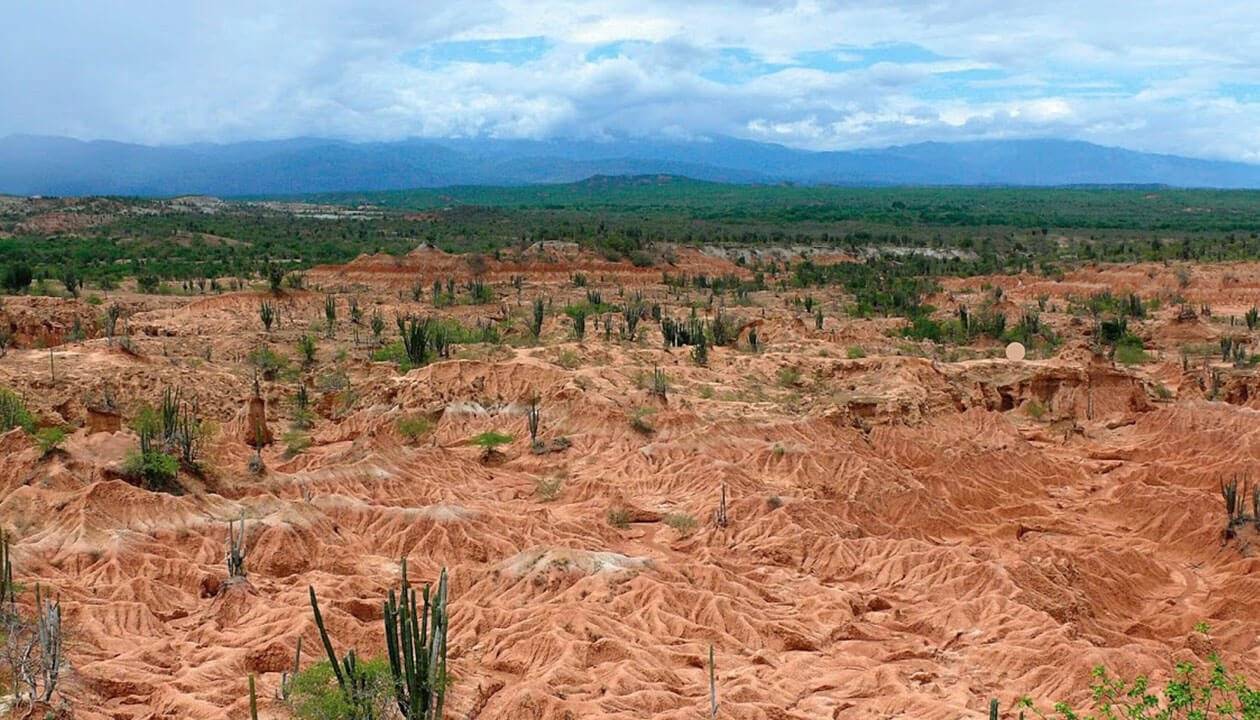 A wide view of Colombia’s Tatacoa Desert in the Huila Region shows red, arid land with eroded gullies and sparse cacti, while a green strip of vegetation and cloudy mountains appear in the distant background.