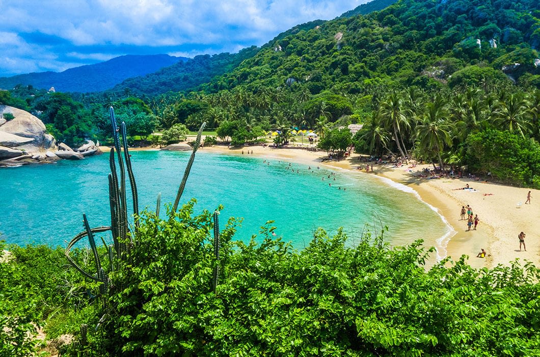 A scenic tropical beach, one of the top Colombia attractions, with turquoise water, lush green hills, palm trees, and people swimming and relaxing on the sandy shore under a partly cloudy sky. Tall cacti are in the foreground.