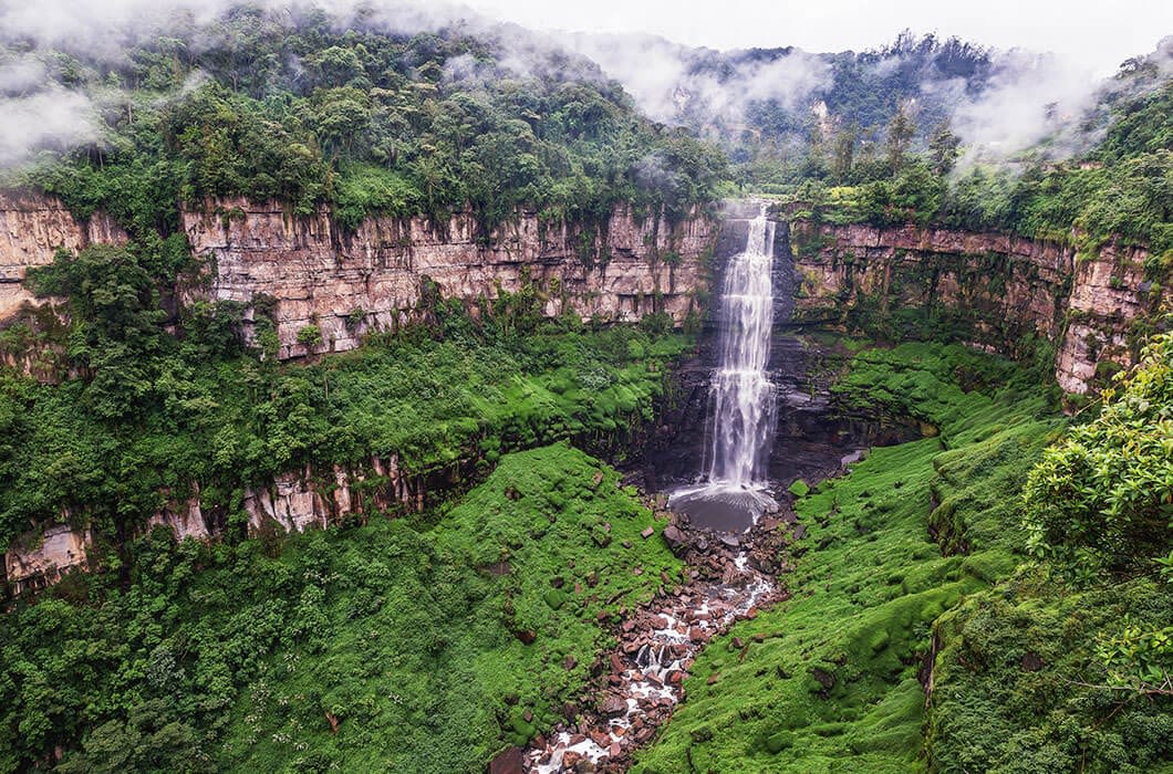 A tall waterfall cascades down a lush, green cliff into a circular pool below, surrounded by dense forest and mist—one of the stunning Colombia attractions perfect for your Colombia travel itinerary.