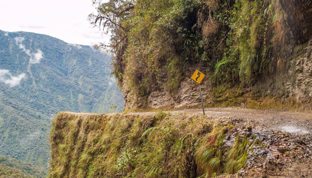 A narrow, winding dirt road clings to the edge of a steep mountain cliff with a sharp curve sign—one of South America's dangerous roads. Dense green forest covers the mountainside, and mist lingers in the background.