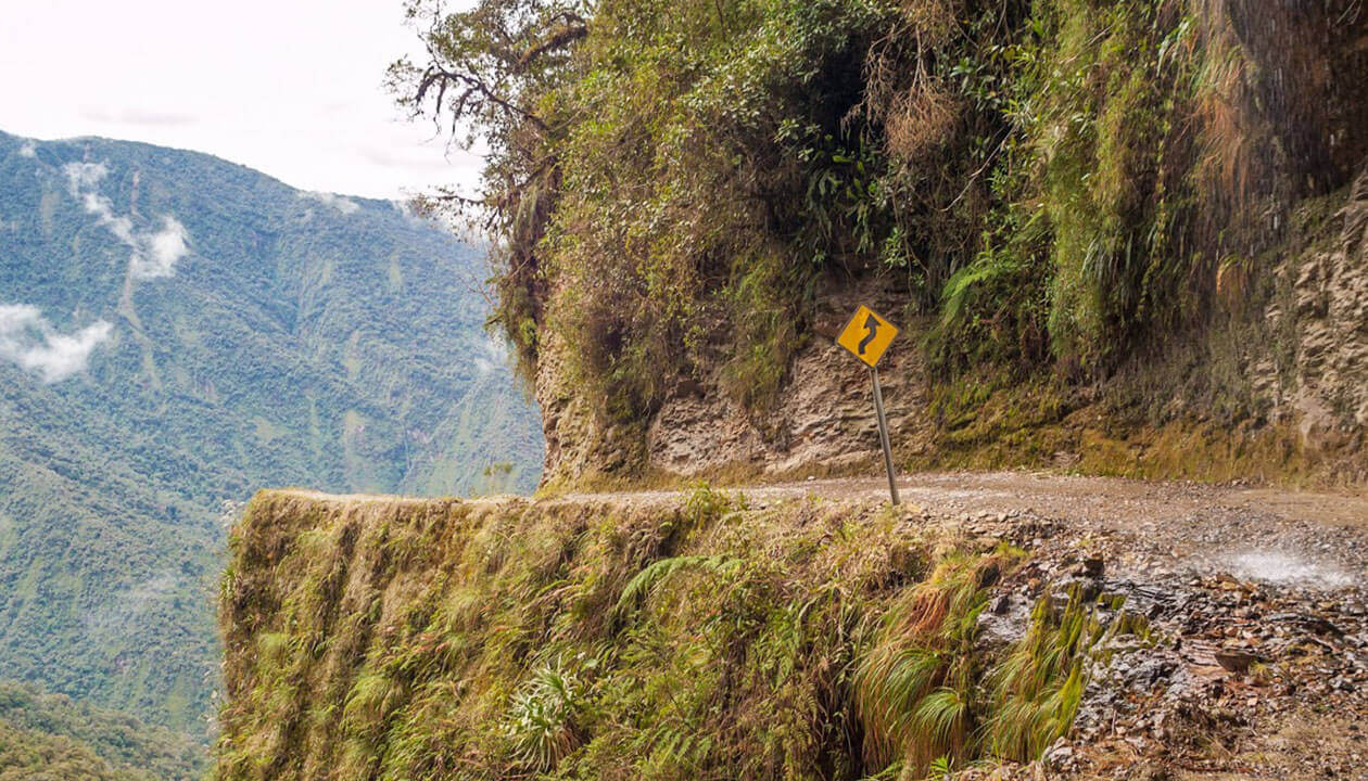 A narrow, winding dirt road clings to the edge of a steep mountain cliff with a sharp curve sign—one of South America's dangerous roads. Dense green forest covers the mountainside, and mist lingers in the background.