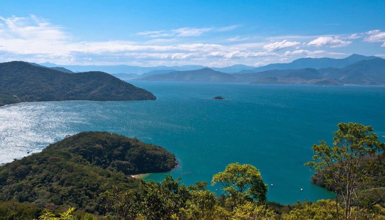 A scenic view of a turquoise bay on one of the best islands in South America, surrounded by lush green hills and distant mountains. Dense forest lines the shorelines while calm water reflects sunlight under a blue sky with scattered clouds.