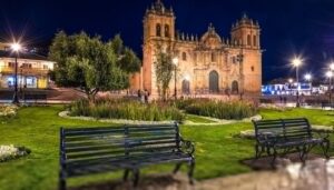 Night view of a historic cathedral with ornate architecture, seen behind a green park with benches, flowers, and trees. Bright streetlights illuminate the scene—one of the must-see Cusco attractions for evening strolls and unforgettable photos.