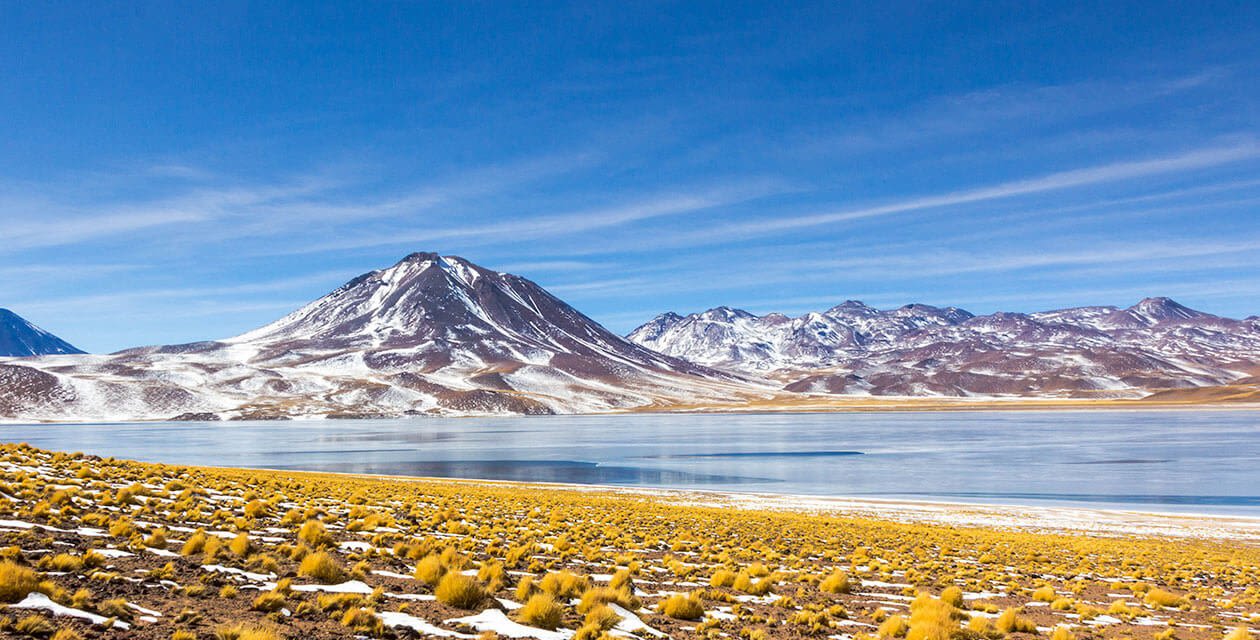 A snow-dusted mountain range stands behind a clear blue lake in South America, with yellow grass tufts scattered across a dry, rocky plain under a bright blue sky—an inviting scene in one of the continent’s safest countries for travel.