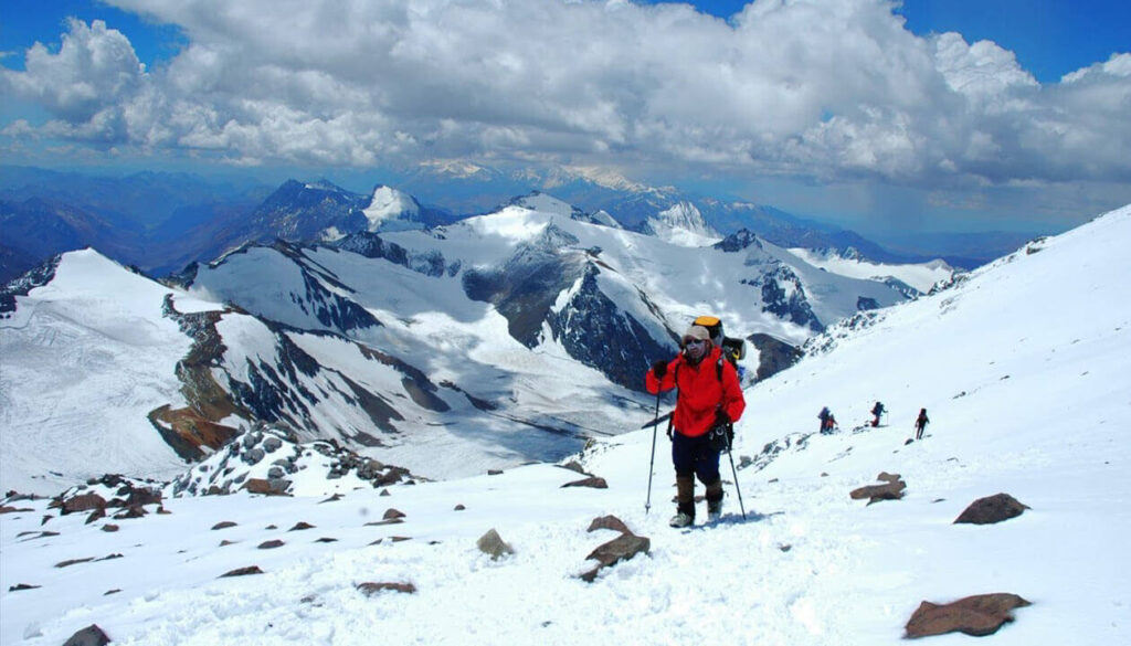 A group of climbers in bright jackets ascend a snowy mountain slope near some of the best ski resorts Argentina offers, surrounded by rugged, snow-covered peaks under a partly cloudy sky.