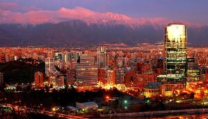 A vibrant cityscape at sunset with modern high-rise buildings and lights glowing, set against snow-capped mountains under a colorful sky—perfect inspiration for Argentina and Chile tours in South America.