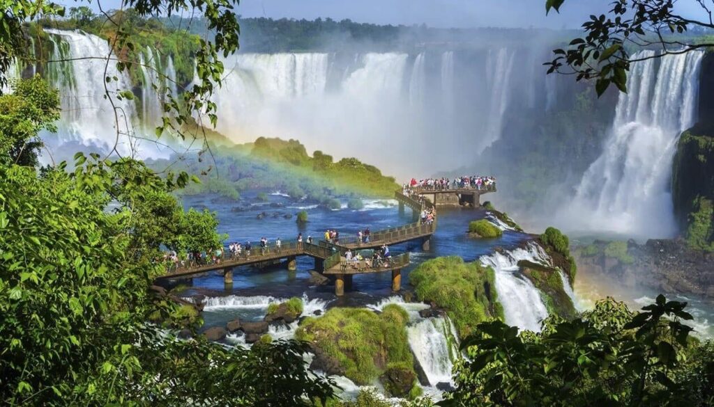 Tourists stand on a boardwalk over rapids, surrounded by lush greenery and tall waterfalls, with a rainbow in the mist at Iguazu Falls—one of the best places to visit in South America for travel enthusiasts.