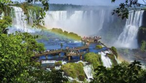 Tourists stand on a boardwalk over rapids, surrounded by lush greenery and tall waterfalls, with a rainbow in the mist at Iguazu Falls—one of the best places to visit in South America for travel enthusiasts.