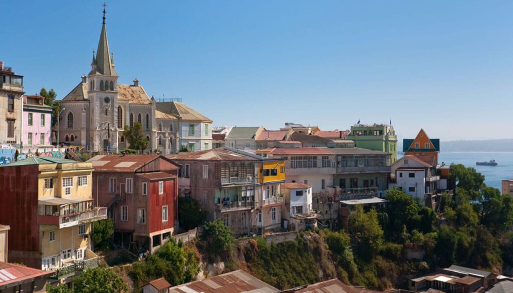 Colorful hillside houses and a church overlook the ocean in Valparaíso, one of the top rated attractions Chile offers. A ship crosses the water as lush greenery surrounds these iconic sights, making Valparaíso a gem of Chile tourism.
