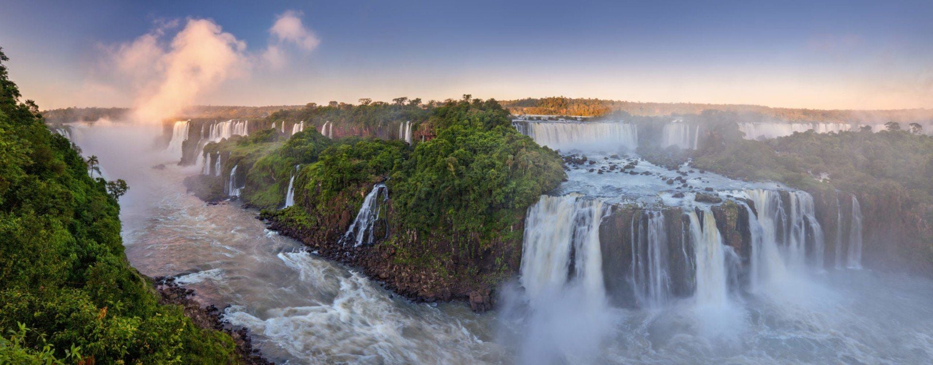 Panoramic view of Iguazu Falls with lush green vegetation, multiple cascading waterfalls, and mist rising into a blue sky at sunrise or sunset—your ultimate travel guide to this breathtaking destination.